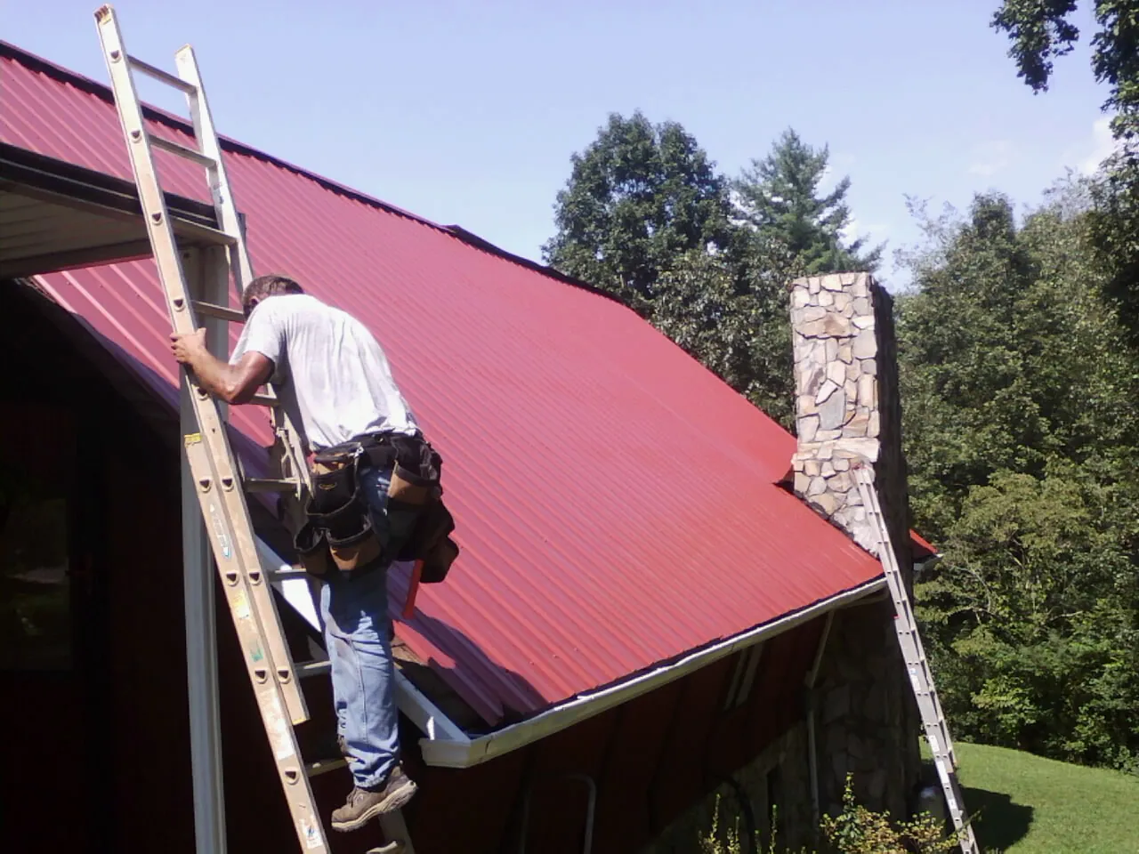 Roofer on a ladder beside a red metal roof and stone chimney — Soffit & Fascia Repair by Roofing Brothers in Sultan